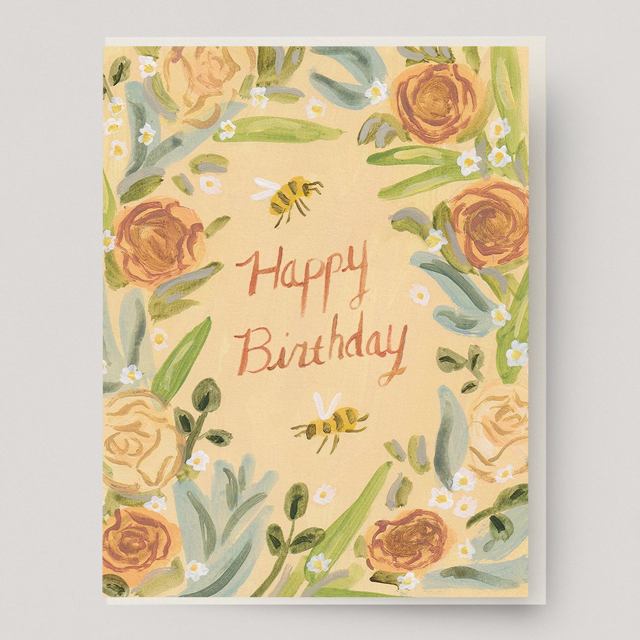 A peach colored greeting card with a wreath of roses, honey bees, daisies and the text "happy birthday" in the center. Hand illustrated by artist Katherine Lewis. Made by Ingrid Press card and gift company, ethically printed in the USA.