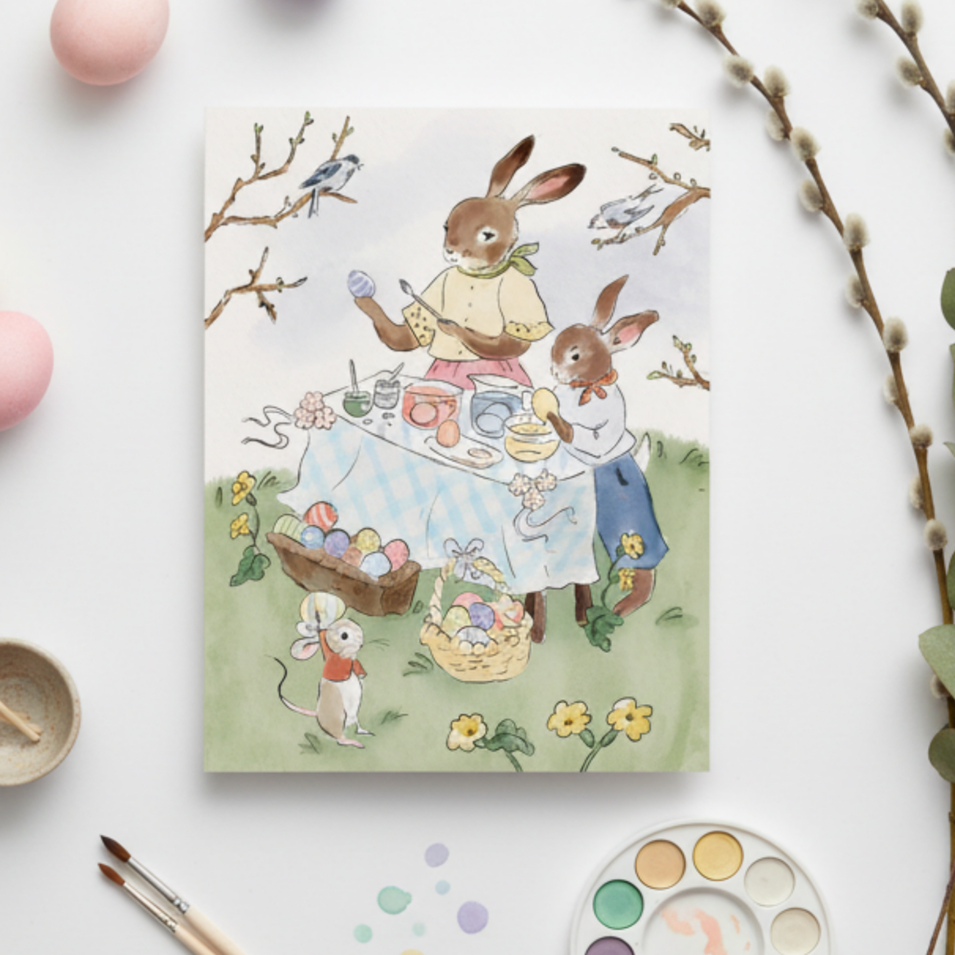 Easter-themed card with rabbits and eggs, surrounded by paintbrushes and pastel colors on a white background.