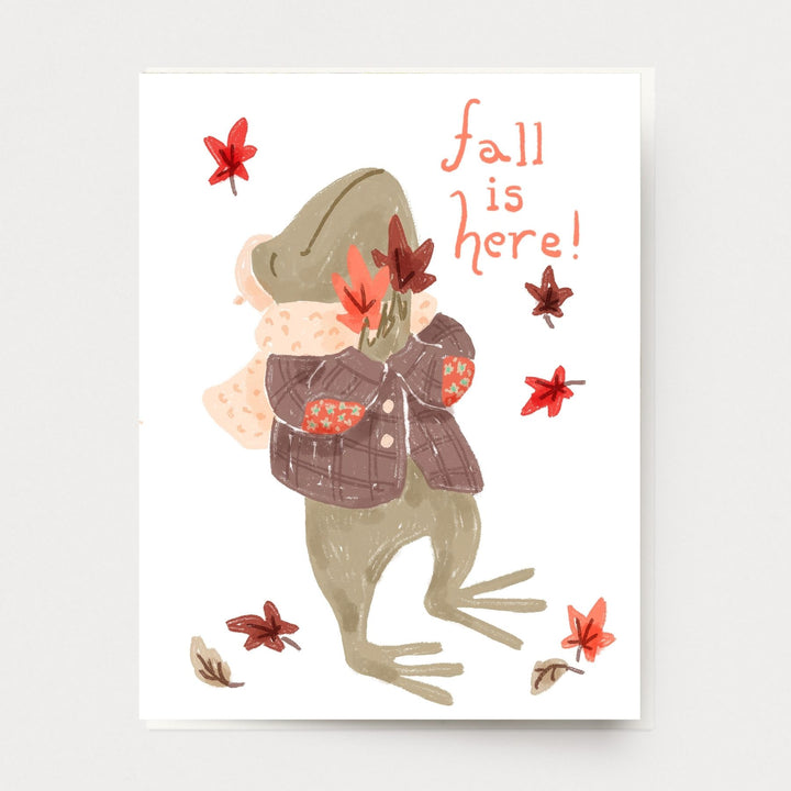 A cute fall card of a frog dressed in a plaid jacket and knitted scarf hugging autumn leaves