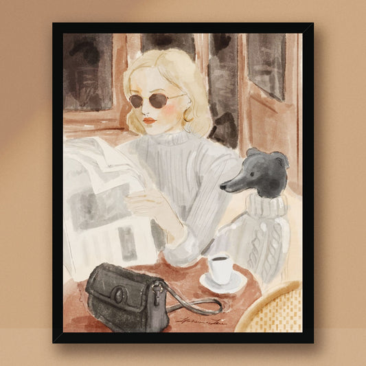 A framed art print featuring a watercolor-style illustration of a blonde woman reading a newspaper, wearing round sunglasses and a light gray sweater, with a black dog and a handbag on a table in the foreground. The print is displayed against a warm beige background.