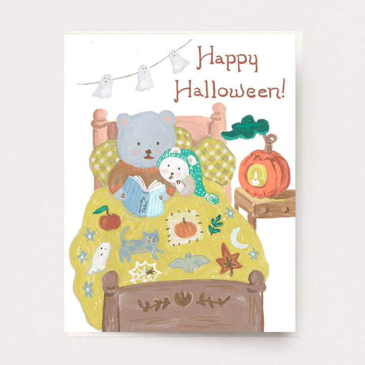 Greeting card of a cute bear reading a book to a young bear cub on a bed with Halloween decorations.