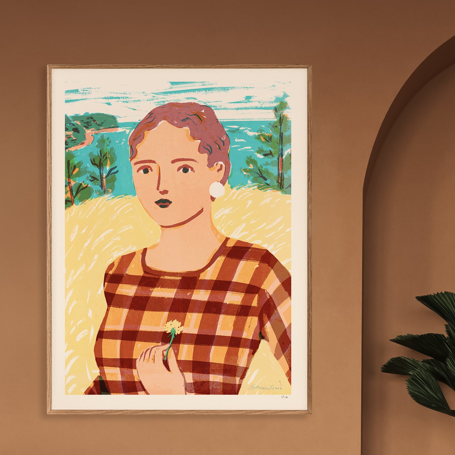 A framed art print featuring a stylized portrait of a woman in a brown and orange plaid top, set against a yellow field and blue-green landscape. The print is displayed on a warm brown wall next to an arched doorway and a green plant.