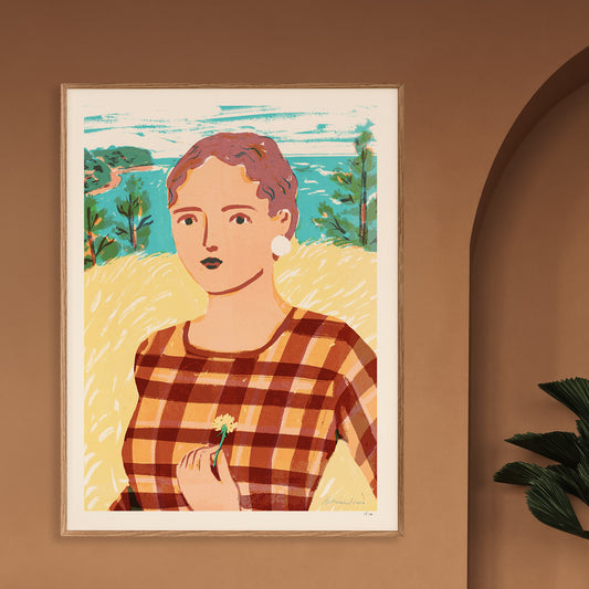 A framed art print featuring a stylized portrait of a woman in a brown and orange plaid top, set against a yellow field and blue-green landscape. The print is displayed on a warm brown wall next to an arched doorway and a green plant.
