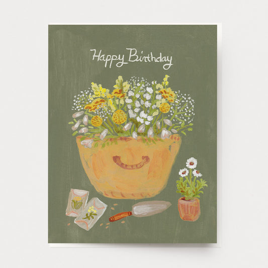 A birthday greeting card with a basket of white and yellow flowers, packets of seeds and a garden trowel, with the words "Happy Birthday" at the top. Hand illustrated by Mendocino Coast artist Katherine Lewis. Made by Ingrid Press card and gift company, ethically printed in the USA.