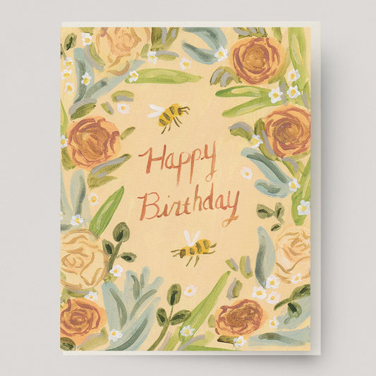 A peach colored greeting card with a wreath of roses, honey bees, daisies and the text "happy birthday" in the center.  Hand illustrated by artist Katherine Lewis. Made by Ingrid Press card and gift company, ethically printed in the USA.