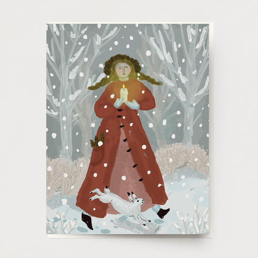 A greeting card of a winter Holiday scene with a woman in a long red coat, long braids and holly berry crown, singing carols, holding a candle, accompanied by a white rabbit. Snow is falling all around.  Hand illustrated and made by Ingrid Press card and gift company, ethically printed in the USA.