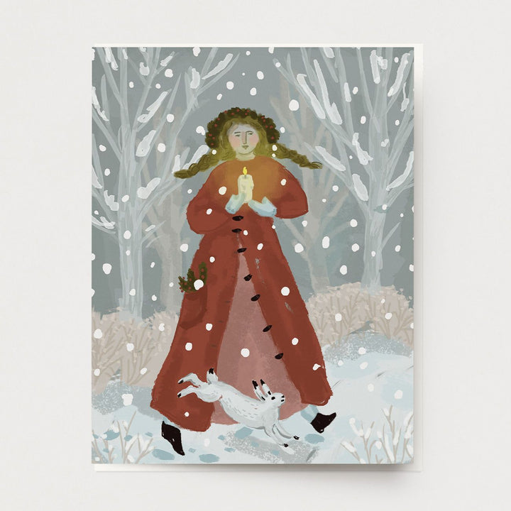 A greeting card of a winter Holiday scene with a woman in a long red coat, long braids and holly berry crown, singing carols, holding a candle, accompanied by a white rabbit. Snow is falling all around.  Hand illustrated and made by Ingrid Press card and gift company, ethically printed in the USA.